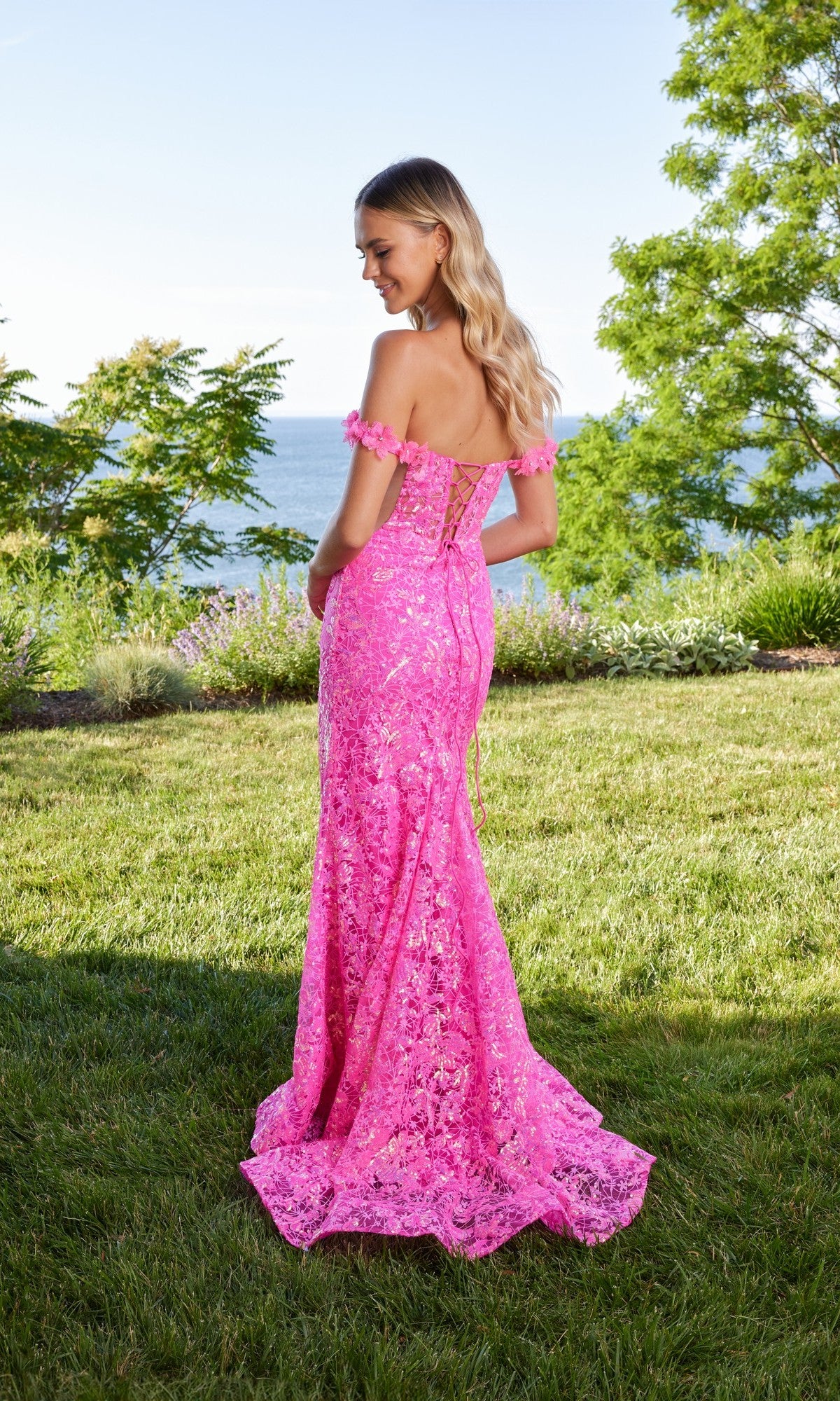 A woman stands outdoors on grass in a vibrant hot-pink Morilee 49188 prom dress—an off-the-shoulder lace gown—with her back to the camera; trees and water create a serene background.
